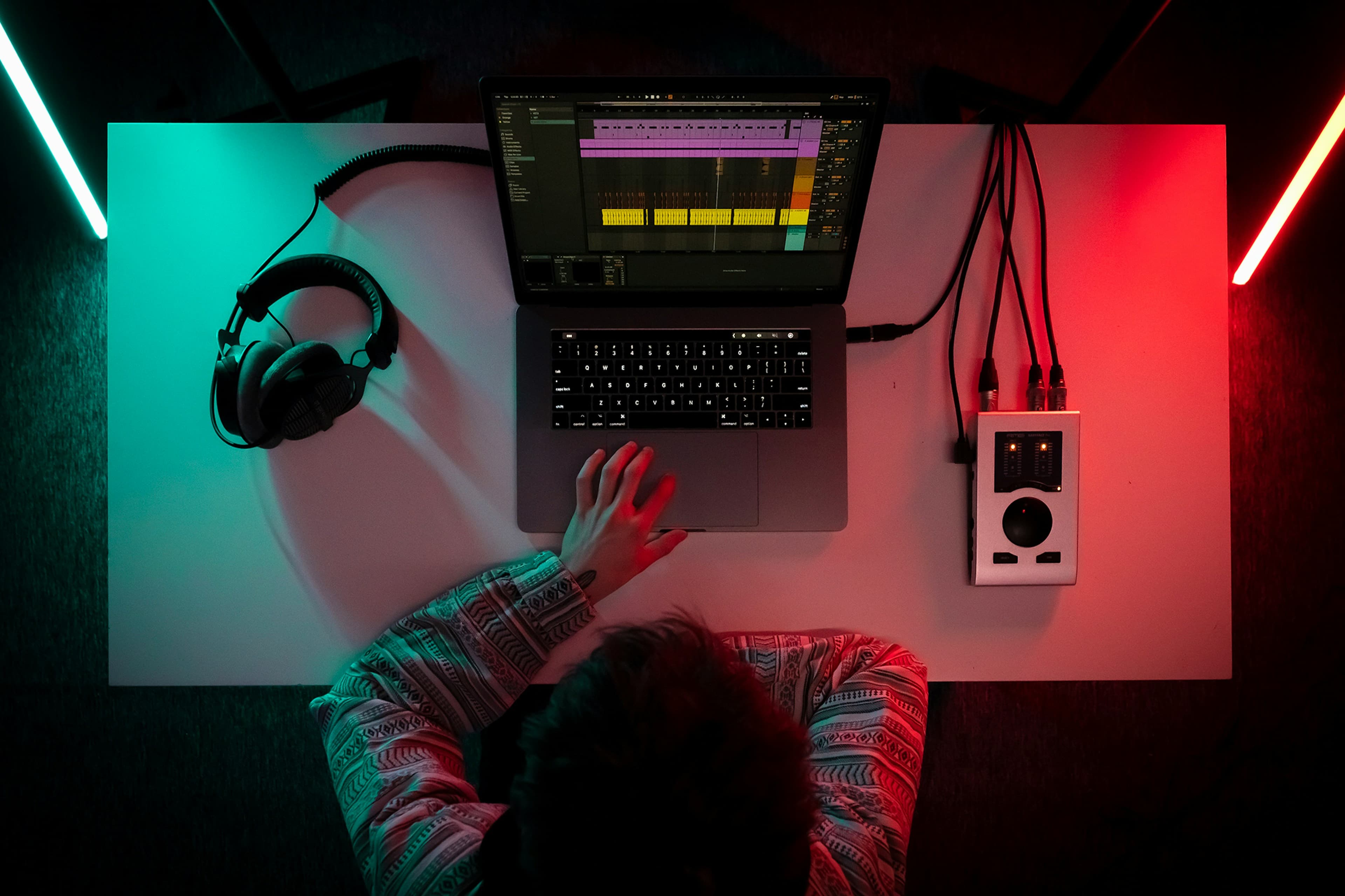 Overhead view of a music production setup — laptop, headphones, and audio interface under colored studio light.